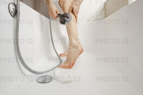 A person is standing in a stylish bathtub while washing her legs with a handheld shower head. Water is flowing freely as part of a soothing self-care routine in a contemporary bathroom