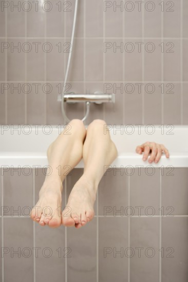 A pair of bare legs rests over the edge of a bathtub, with feet comfortably hanging. A hand is seen reaching out from behind, creating a relaxed and serene atmosphere within the bathroom