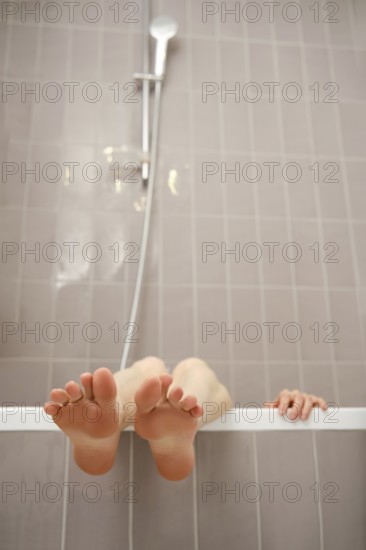 Feet sticking out over the edge of bathtub. The modern bathroom features light-colored tiles, and a showerhead