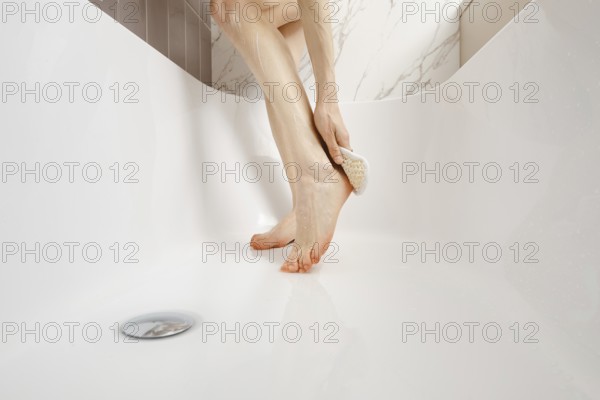 A person enjoys a moment of self-care, using a foot scrub in a clean, white bathtub. Natural light illuminates the serene bathroom, enhancing the tranquil atmosphere of the wellness activity