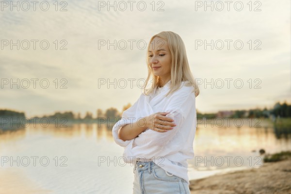 A young woman stands by the calm lakeside at sunset, wrapped in a cozy white shirt and denim jeans, reflecting on the tranquil waters and the colorful sky, embodying peace and serenity