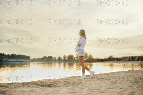 A young woman explores a sandy beach as the sun sets over a calm lake. The scenery features beautiful reflections on the water, warm colors in the sky, and a serene atmosphere among the trees