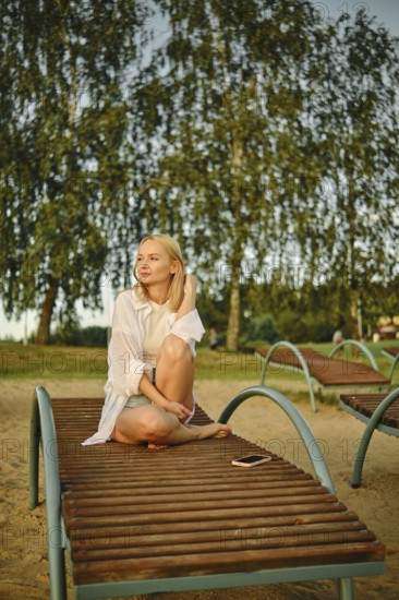 A woman sits comfortably on a lounge chair at the beach as the warm sunlight illuminates her surroundings. Trees sway gently in the breeze, creating a peaceful atmosphere during the evening