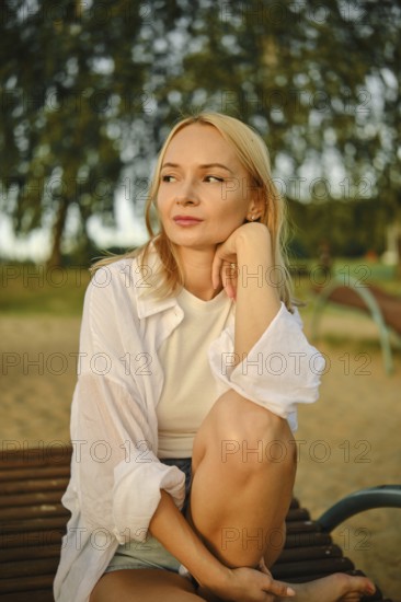 A young woman with blonde hair rests her chin on her hand while sitting on a bench in a park. Sunlight filters through trees, creating a serene atmosphere during the late afternoon hours
