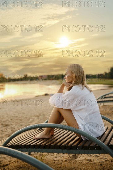A woman sits pensively on a wooden lounge chair by the lakeside as the sun sets. She gazes at the water, reflecting golden hues from the fading light, surrounded by a serene atmosphere