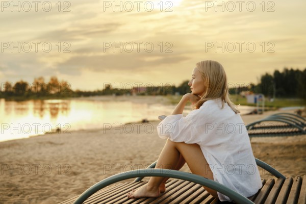 A woman sits on a wooden bench by the shore, gazing at a stunning sunset over the water. The soft golden light reflects on the calm surface, enhancing the peaceful atmosphere of the evening