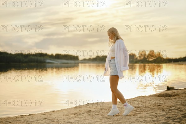 A young woman strolls on the sandy beach at sunset, with the golden light reflecting on the calm water. She is dressed in a casual outfit, enjoying the serene atmosphere and picturesque surroundings