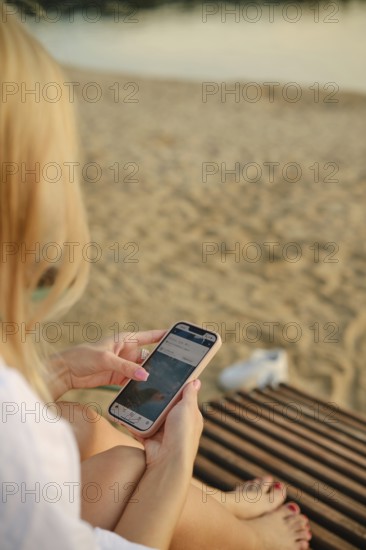 A woman sits on a wooden lounge, absorbed in her smartphone. The sun sets in the background, casting warm colors across the beach, creating a serene and relaxing atmosphere