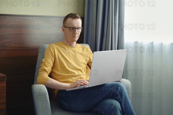A man sits in a comfortable chair, focused on his laptop in a bright room. Soft natural light streams through the curtains as he engages in his tasks, showcasing a productive atmosphere