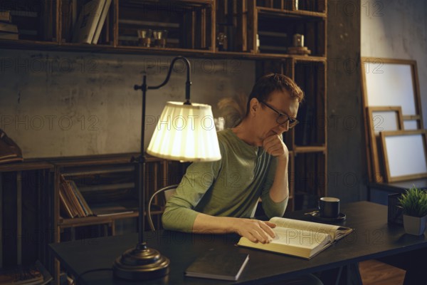 A man sits at a desk in a dimly lit room, engrossed in a book. Warm light from a lamp highlights the pages while a cup rests by his side. Bookshelves filled with books create a calming atmosphere