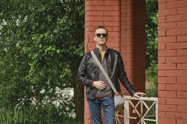 A man stands with a relaxed posture beside vibrant red brick pillars, dressed in a casual outfit featuring sunglasses and a messenger bag