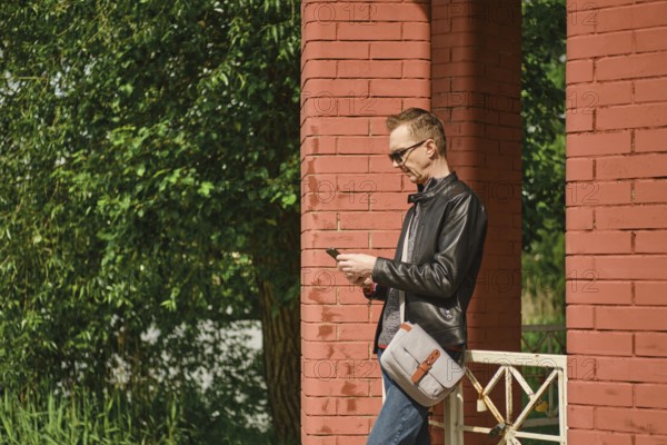 A man wearing sunglasses and a leather jacket stands by red brick columns, attentively using his smartphone. Lush green foliage surrounds the structure, indicating a pleasant sunny day