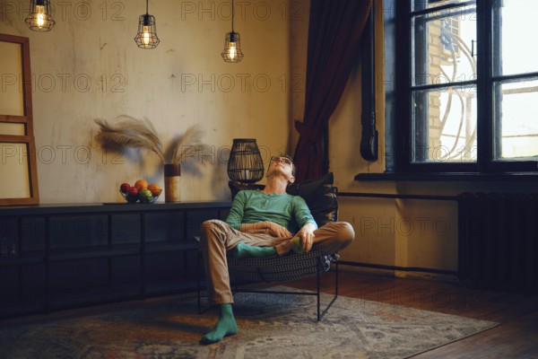A man relaxes in a modern chair in a well-lit living space filled with warm tones. Sunlight streams through a large window, highlighting unique decor elements and a bowl of fruit nearby
