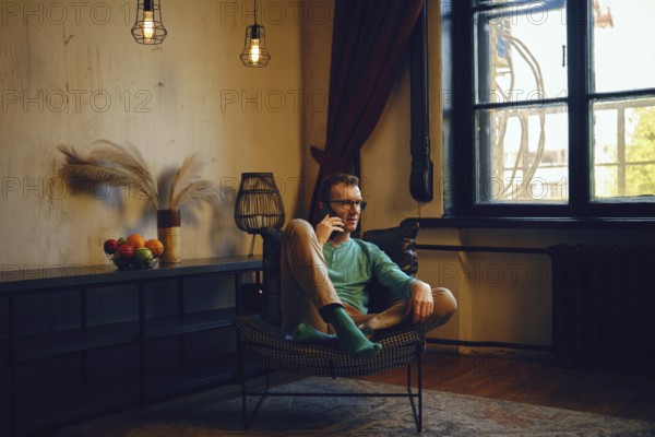 A man dressed in a green long-sleeve shirt and light pants relaxes in a chair, engaging in a phone conversation. The room features warm lighting, a window, and a basket of fruits nearby