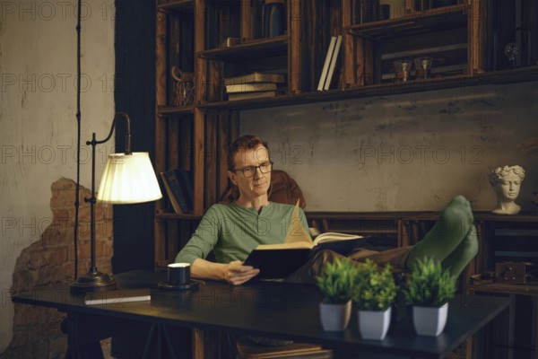 A middle-aged man relaxes with his feet up on a desk in a home office, reading a book. Natural light illuminates the cozy space, filled with books and decorative elements