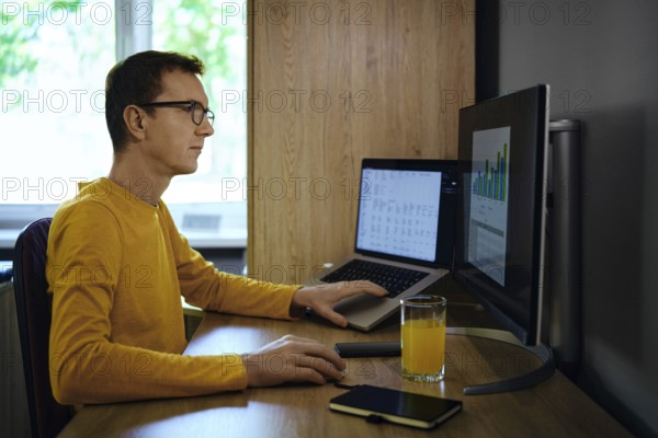 A man wearing a yellow shirt sits focused at a desk, using a laptop and a monitor. He analyses data and predicts the growth of his investments in real estate