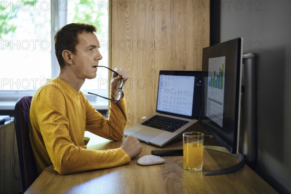 A man in a yellow shirt sits at a desk in his home office, studying data on his computer and bites the rim of his glasses