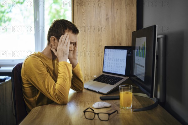 A middle-aged man sits at a wooden desk holding his head in frustration. Two computer screens display data of loan interest rate