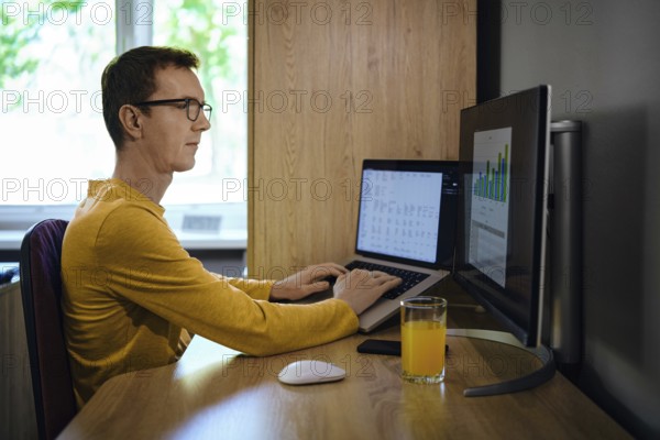 A middle-aged man focused on his work sits at a wooden desk with two computer screens. He enters various data and looks at the chart, analyzing and predicting various scenarios
