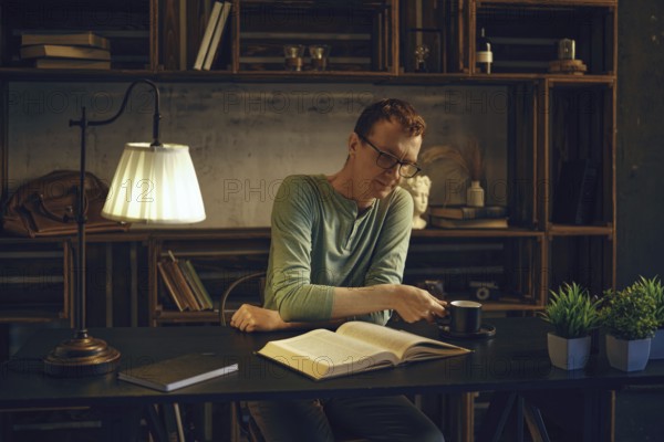 A man enjoys a quiet evening in a rustic home office, reading a book while sipping herbal tea. The warm lighting creates a cozy atmosphere, with plants and shelves in the background adding charm
