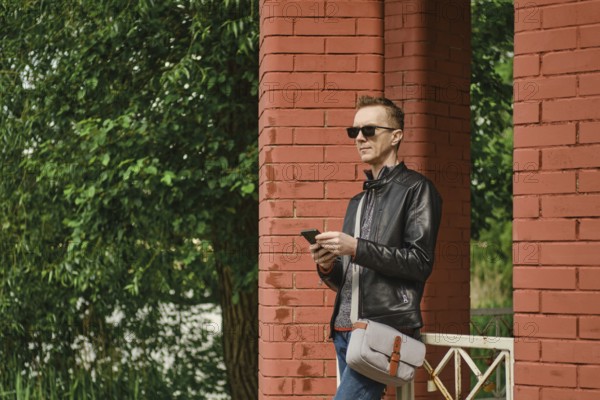 A man dressed in a leather jacket and sunglasses stands by red brick columns. He holds a smartphone and looking for an approaching taxi