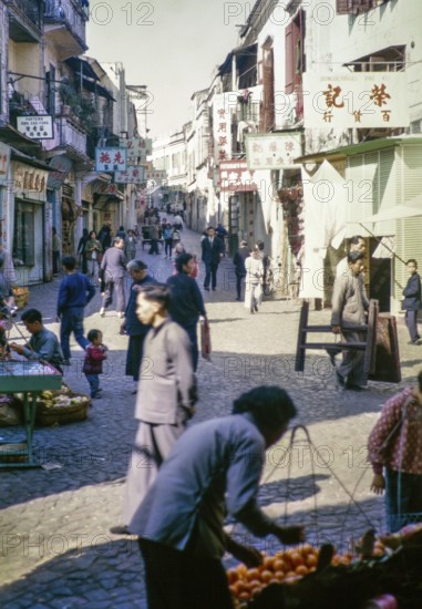 Busy shopping street Cantonese shop signs, with people walking traders with fruit stalls, Macau, Asia 1964