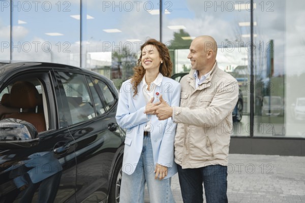 A couple is happily engaged in a conversation while selecting a new car at a dealership. The woman smiles as she interacts with her partner, who gestures toward the vehicle, suggesting its features