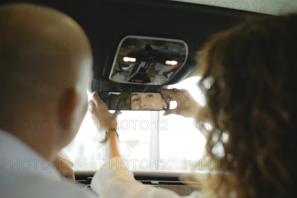 The view from the rear seat of the car is a couple of sitting in front. A woman sets up a rear view mirror and smiles