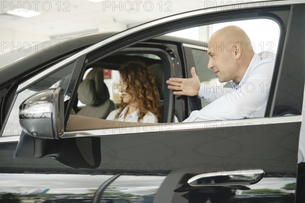 A woman evaluates a car while seated inside it at a car dealership. A salesperson gestures towards features, engaging her in conversation