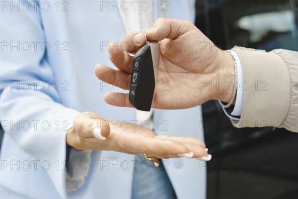 A person hands over car keys to another individual in a parking lot. The transaction takes place under bright sunlight, showcasing the keys prominently as they change hands