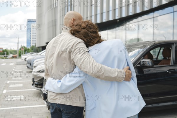 A couple stands affectionately close while considering car options at an automobile dealership. They appear excited as they gaze at the vehicles lined up in the lot under a bright sky