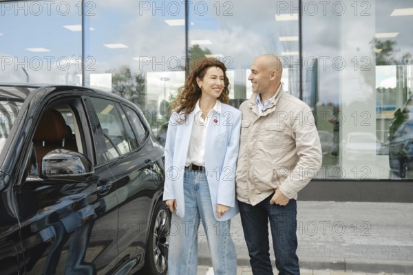 A couple stands beside a sleek black car at a bustling car dealership, engaged in a lively conversation about their choices