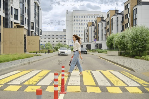A woman with long hair walks across a marked pedestrian crossing in a contemporary urban area. She wears casual clothing and carries a bag