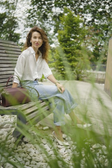 A woman with curly hair relaxes on a wooden bench in a serene park setting, wearing a white shirt and light blue jeans. She has a handbag beside her and enjoys the warm weather amidst lush greenery