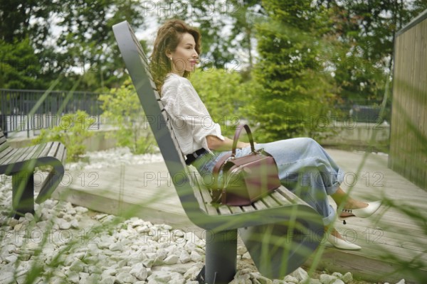 A woman sits comfortably on a bench in a park, surrounded by greenery and decorative stones. She has a stylish handbag and is enjoying the peaceful atmosphere on a bright day