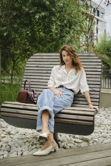 A woman sits comfortably on a unique wooden bench in a modern urban garden. She wears casual clothes with a stylish bag on her side and enjoys the peaceful surroundings filled with greenery