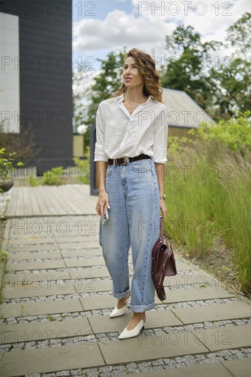 A young woman is walking along a stone path, wearing a white shirt and light blue baggy jeans. She carries a burgundy handbag and checks her phone while surrounded by greenery and modern buildings