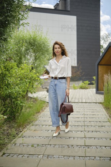 A woman walks gracefully along a garden path surrounded by lush greenery and modern architecture. She wears a stylish outfit, holding her belongings, enjoying a sunny day outdoors