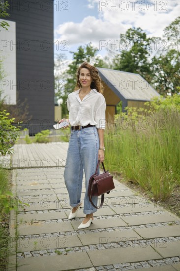 A fashionable woman strolls along a scenic pathway lined with lush plants. She wears a white shirt and high-waisted jeans, carrying a bag and checking her phone while enjoying the outdoors