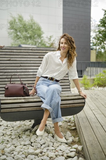 A woman sits comfortably on a modern bench in a lush park, wearing a light button-up shirt and wide-leg jeans. Her stylish bag rests nearby as she enjoys the pleasant weather and surroundings