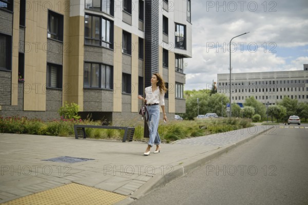 A woman strolls down a sidewalk in a vibrant urban area. She wears a stylish outfit and walks past modern buildings while enjoying the pleasant weather. The street is lively with greenery nearby
