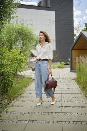 A woman wearing a white shirt and high-waisted jeans strolls through a landscaped path surrounded by greenery