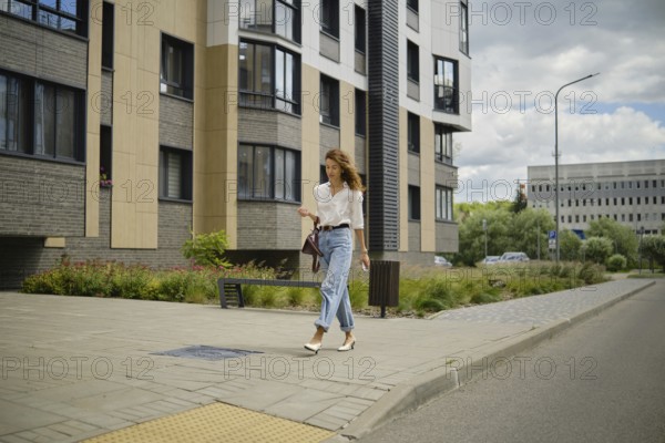 A woman strolls down a city sidewalk on a sunny day. She wears a casual outfit with light jeans and a blouse. Modern buildings and lush plants create a vibrant urban atmosphere