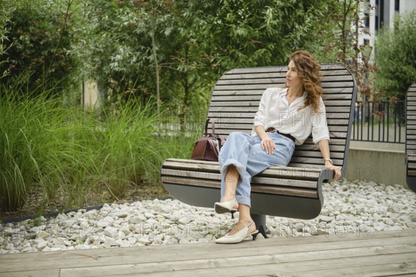 A woman with curly hair is seated on a modern wooden bench in a landscaped urban park. She is wearing a casual outfit, enjoying the surrounding greenery and the warm daylight