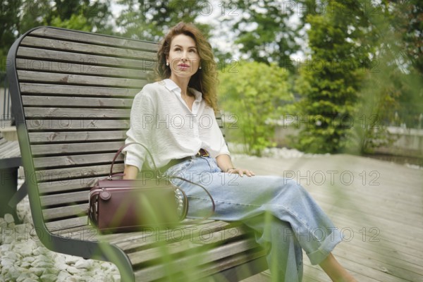 A woman with curly hair sits comfortably on a wooden bench in a lush park. Dressed in a light shirt and high-waisted jeans, she enjoys the tranquil surroundings while placing her handbag beside her