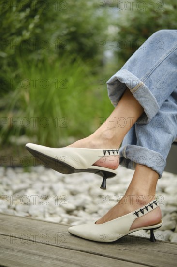 A woman showcases fashionable white slingback heels while sitting outdoors. She wears rolled-up denim jeans, surrounded by lush greenery and pebbles, exuding a casual yet chic vibe