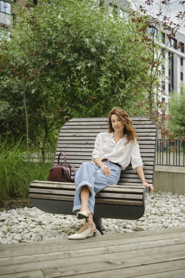 A woman sits comfortably on a contemporary bench in a landscaped area filled with lush green plants. She wears a light blouse and loose-fitting jeans, exuding a relaxed vibe in an urban environment