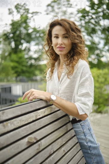 A woman with curly hair stands confidently by a wooden railing in a green park. She wears a light shirt and jeans and enjoys a bright, sunny day surrounded by lush trees and greenery