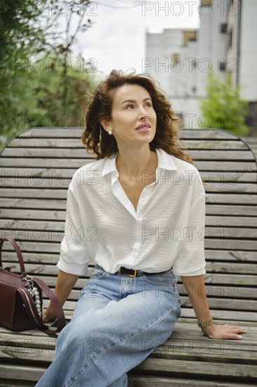 A woman with curly hair sits relaxed on a wooden bench, wearing a white shirt and blue jeans. She looks content while enjoying the warm weather with greenery in the background