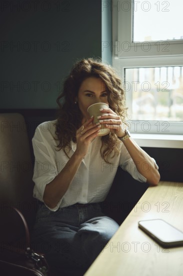 A woman with curly hair sits by a window in a cafe, holding a warm cup of coffee. Sunlight streams in as she enjoys her drink, dressed casually in a white shirt and denim